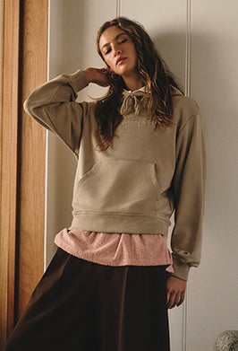A woman stands indoors wearing a beige hoodie over a pink textured shirt and a dark brown skirt, with her hand touching her neck and her eyes partially closed.