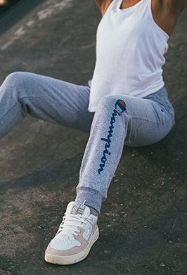 Person sitting on the ground wearing a white tank top, gray Champion sweatpants, and white sneakers. The Champion logo is visible on the side of the pants. The persons face is not shown.